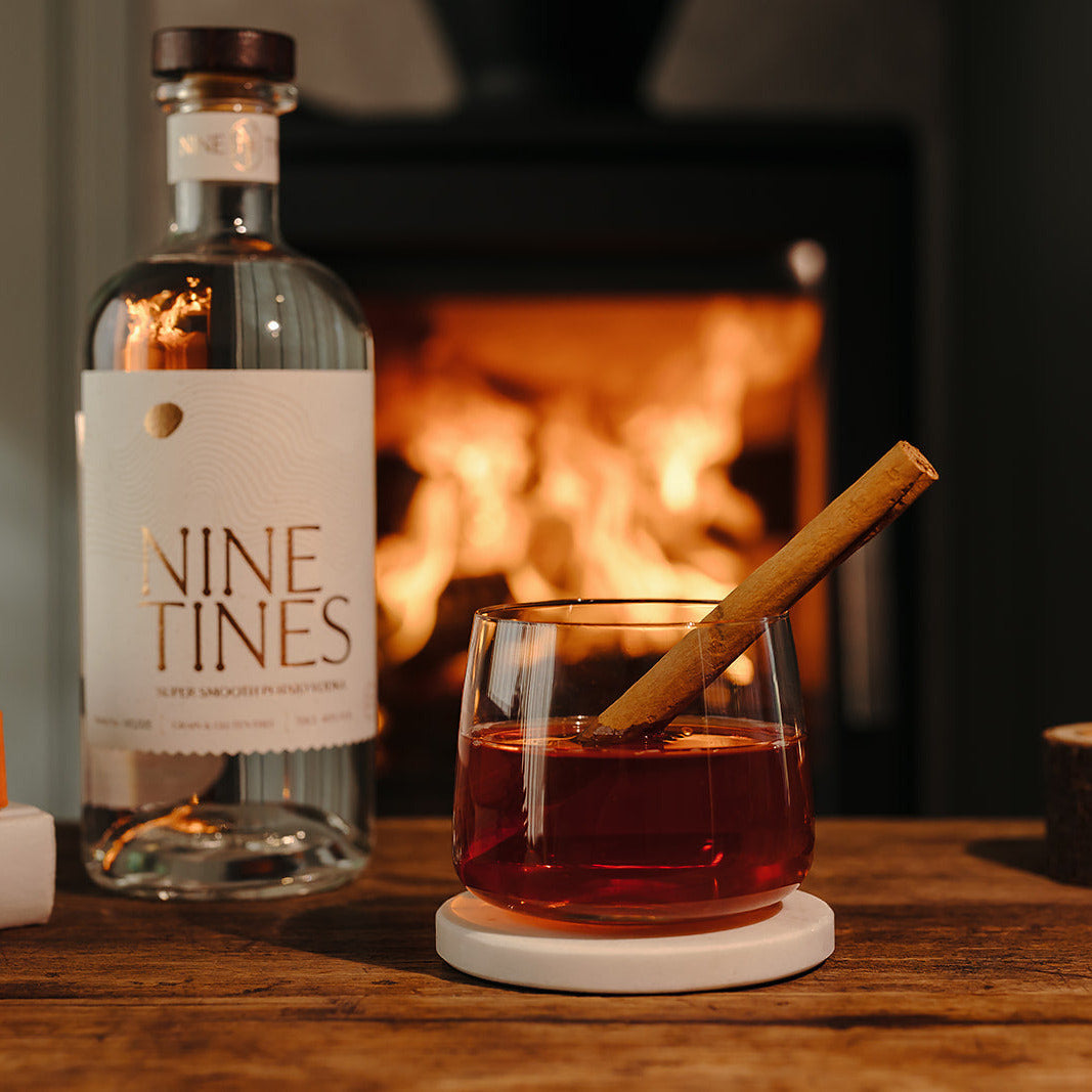 Bottle of Nine Tines vodka with a negroni and a cinnamon stick in front of a fireplace.