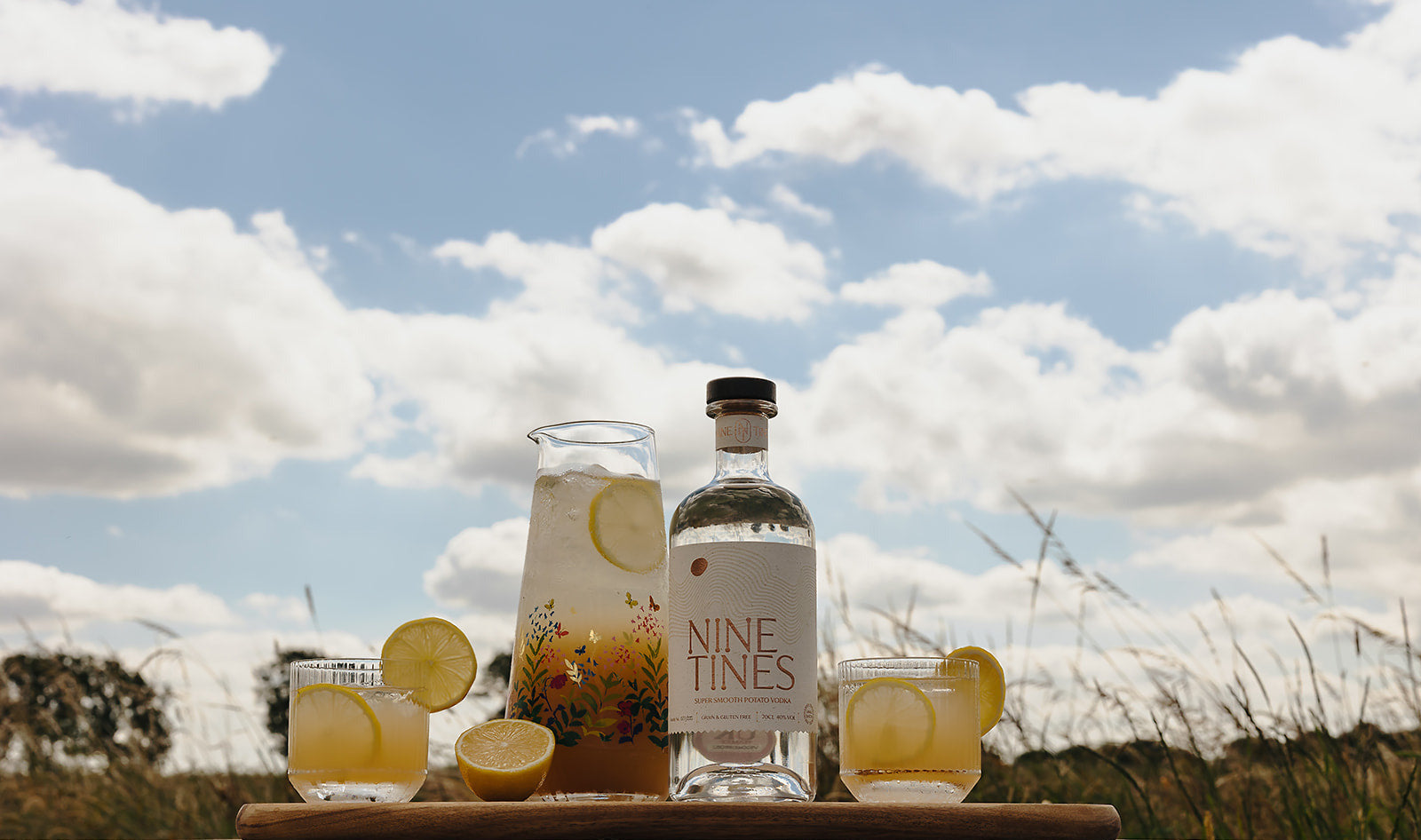 Bottle of Nine Tines gin with glasses of Elderflower Quencher against a summer sky.
